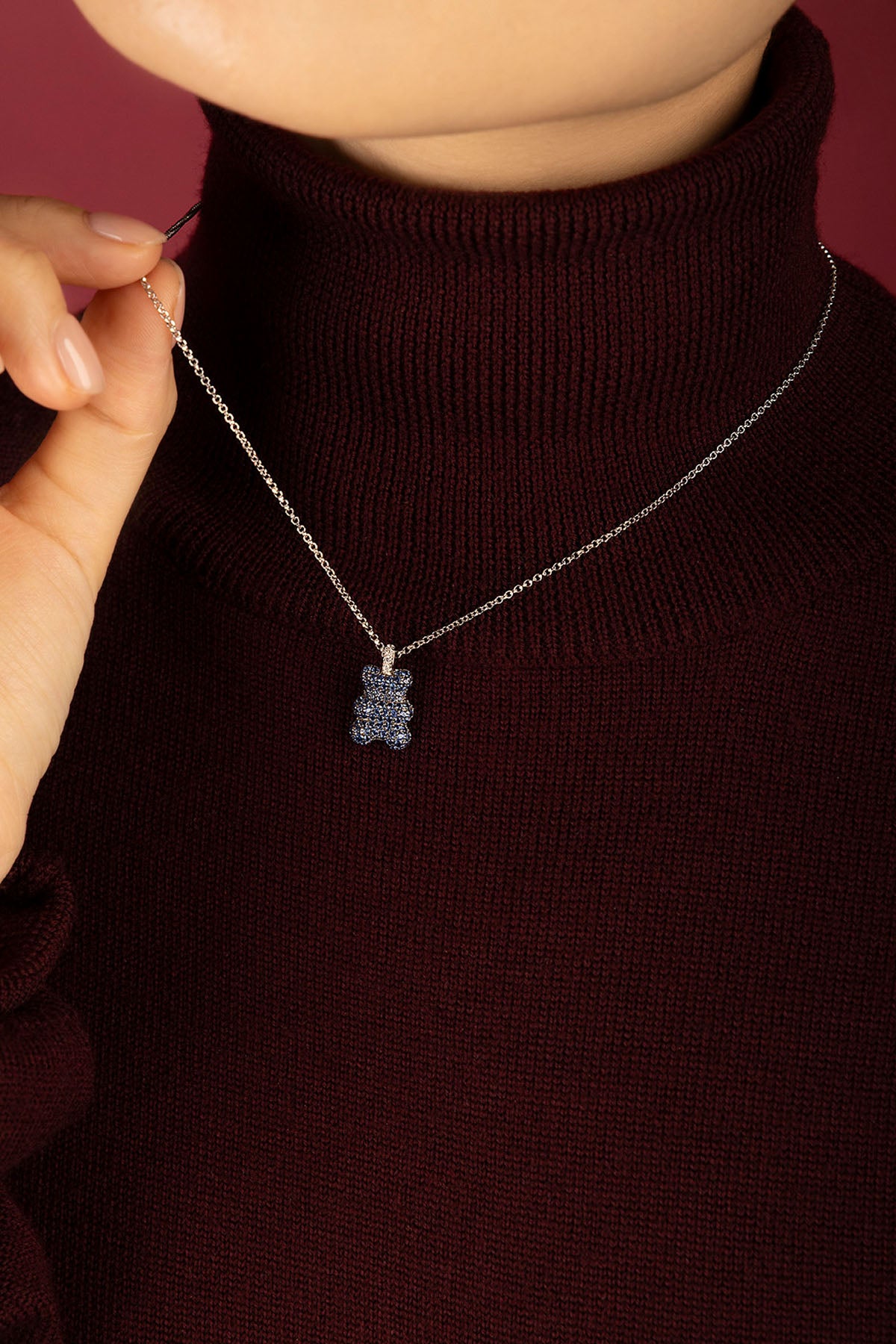 Woman wearing APM Yummy Bear Necklace, a playful blue sparkle against cozy burgundy.