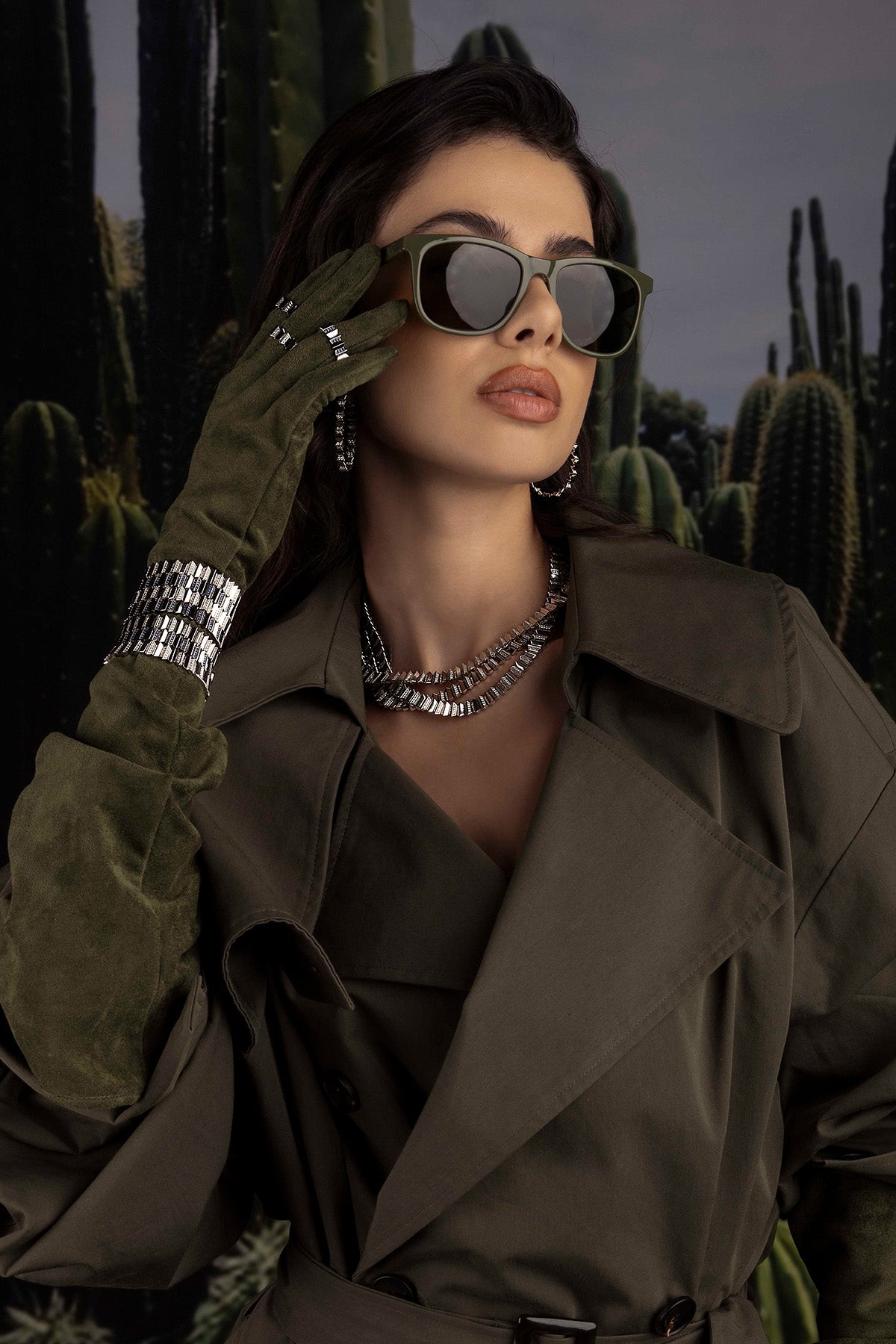Woman in a trench coat, jewelry and sunglasses with cacti in the background
