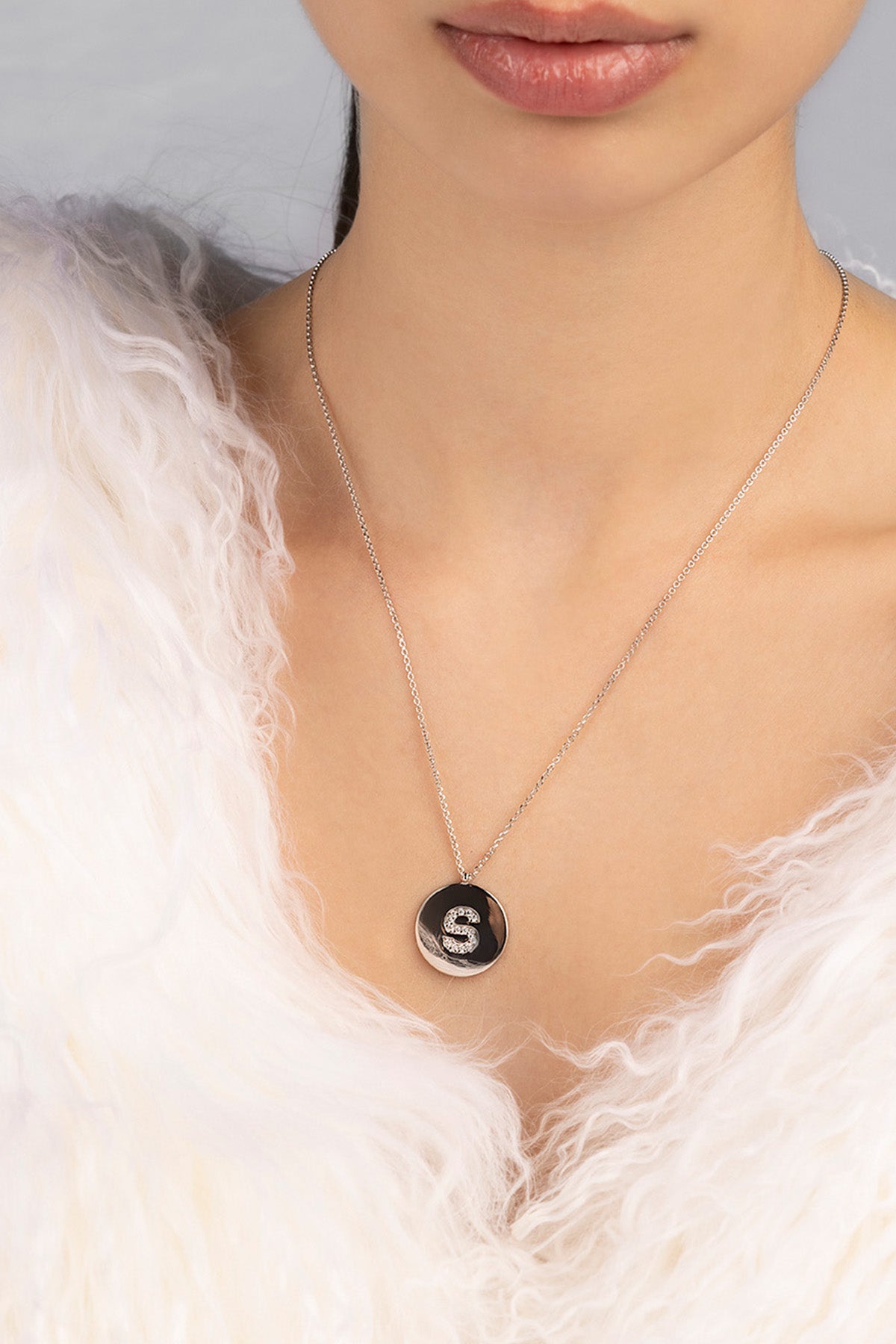 Necklace with a round pendant featuring the letter 'S' worn by a person against a neutral background
