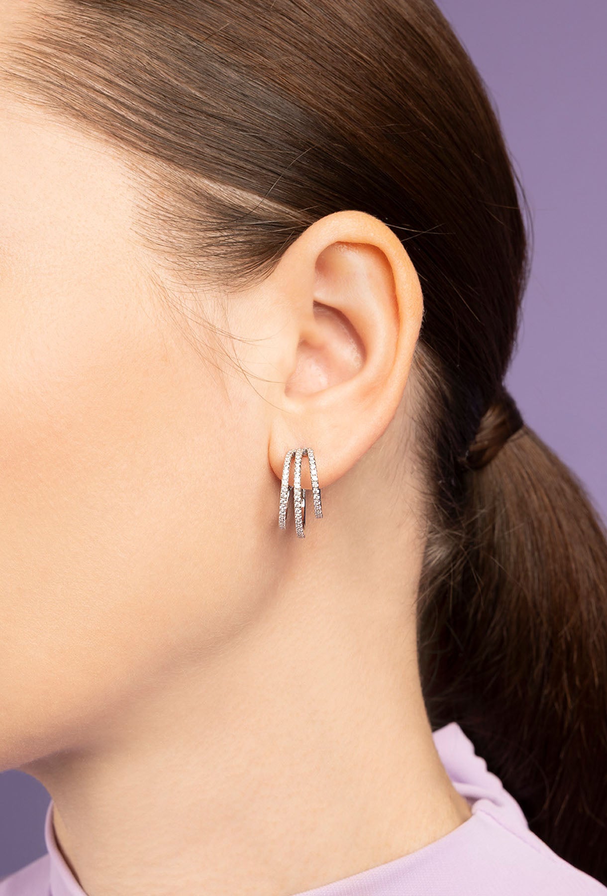 Close-up of a person wearing silver triple hoop earrings with a blurred purple background