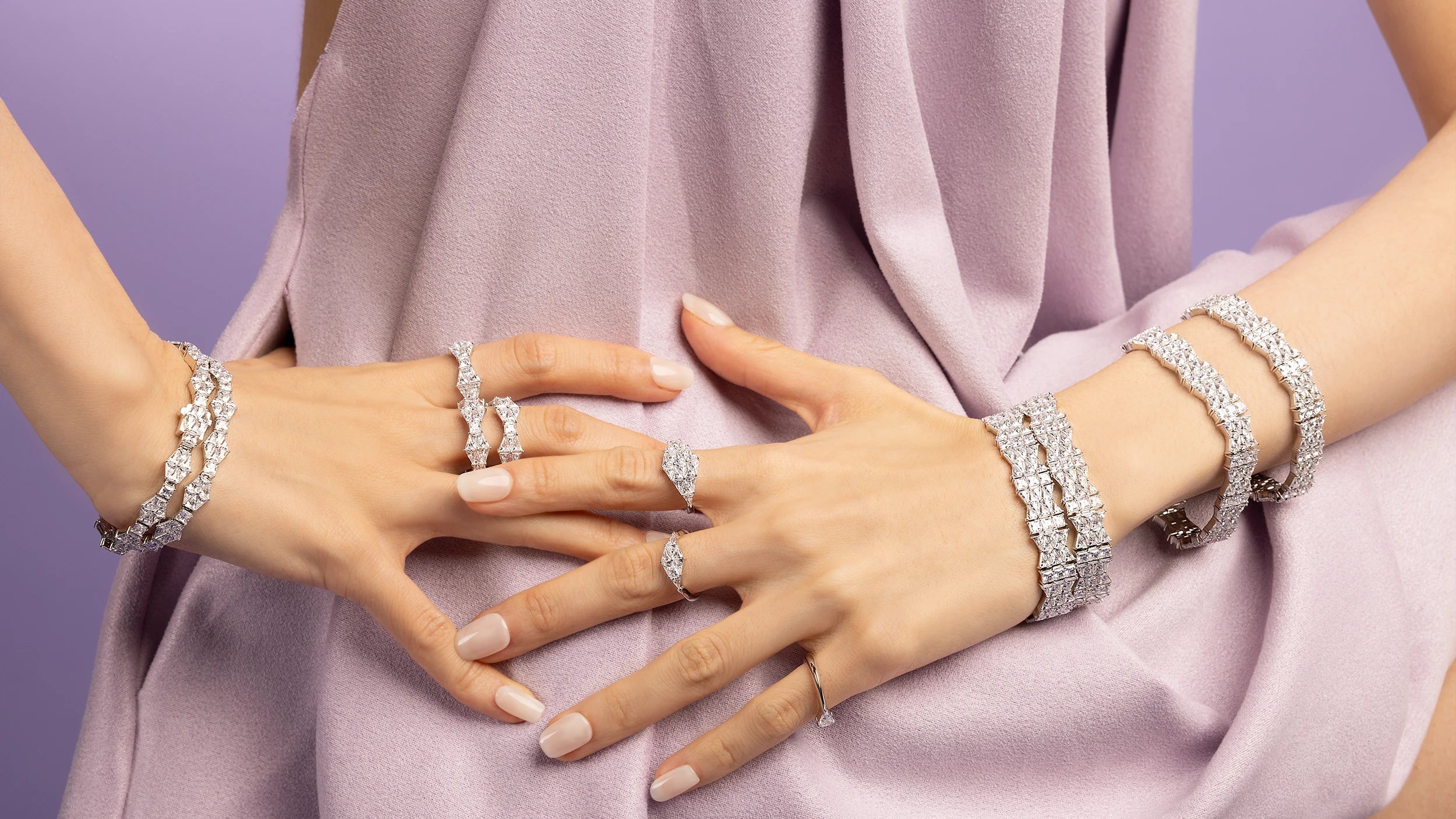 Close-up of hands wearing multiple APM MONACO geometric silver rings and bracelets on a light purple fabric background