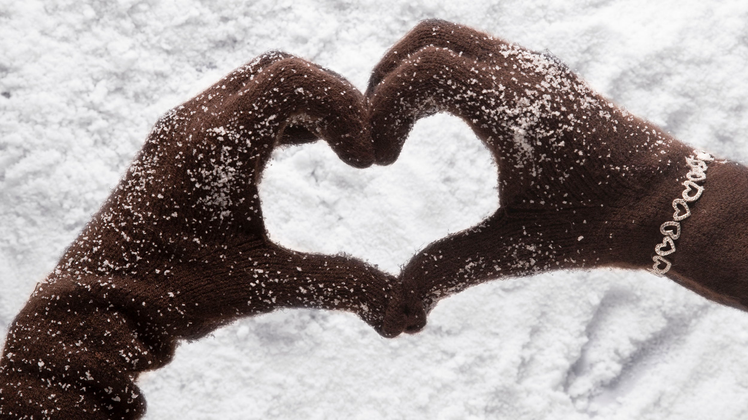 Hands making a heart shape in the snow with APM MONACO silver heart-shaped bracelet
