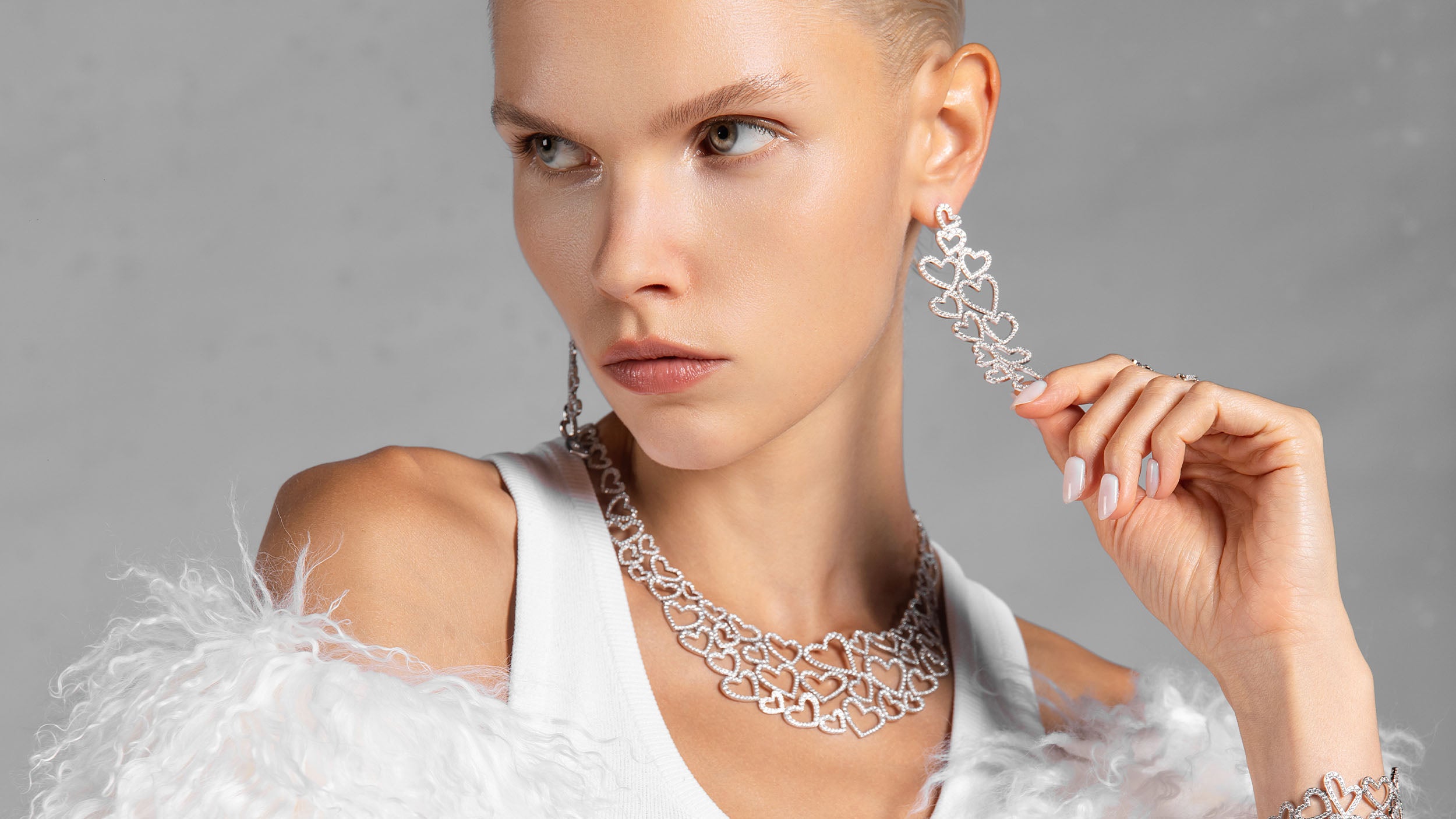 Woman wearing silver heart-shaped jewelry set from APM MONACO, including earrings and a necklace against a gray background