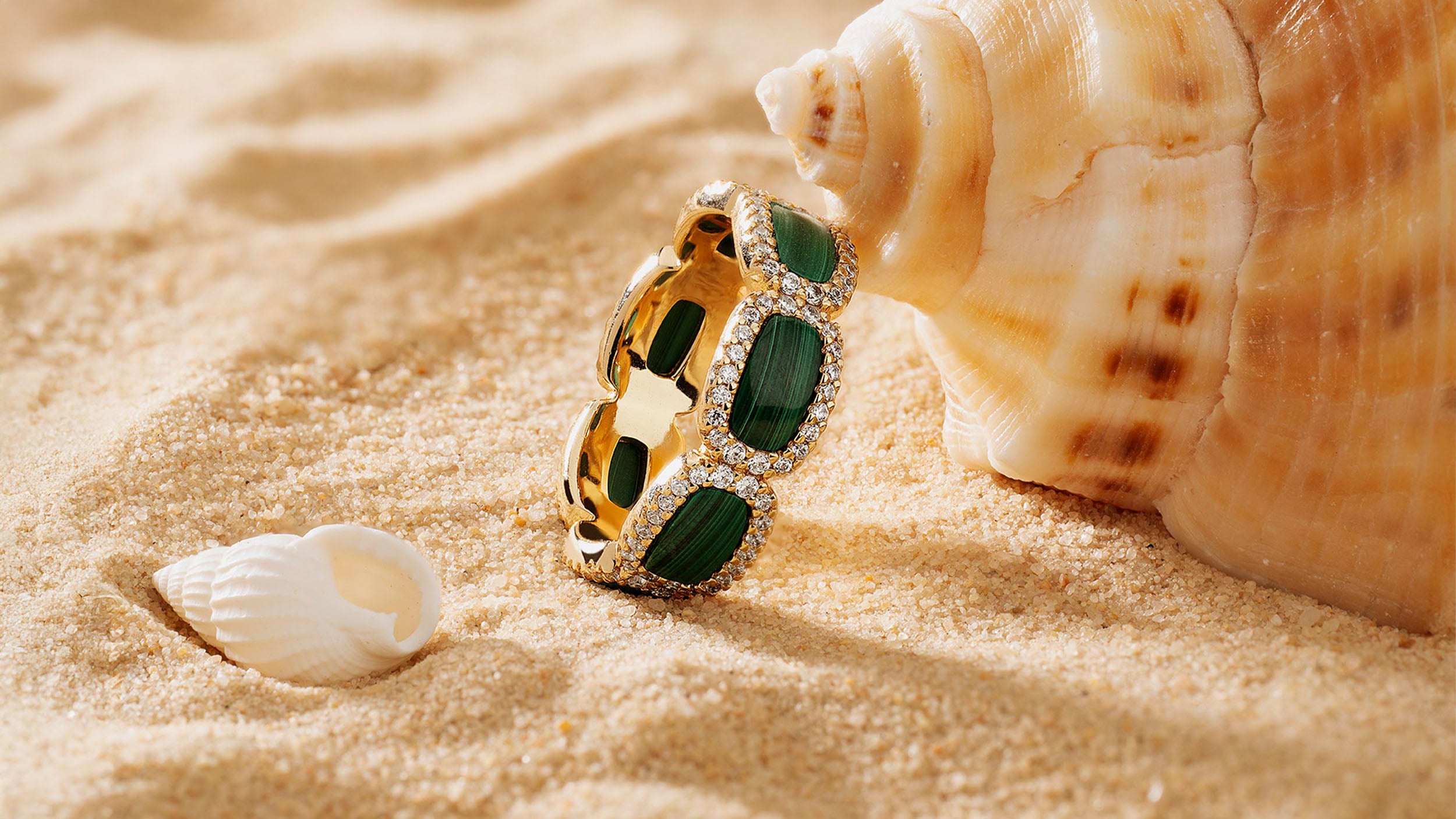 APM MONACO yellow gold ring with green stones on sand next to a shell