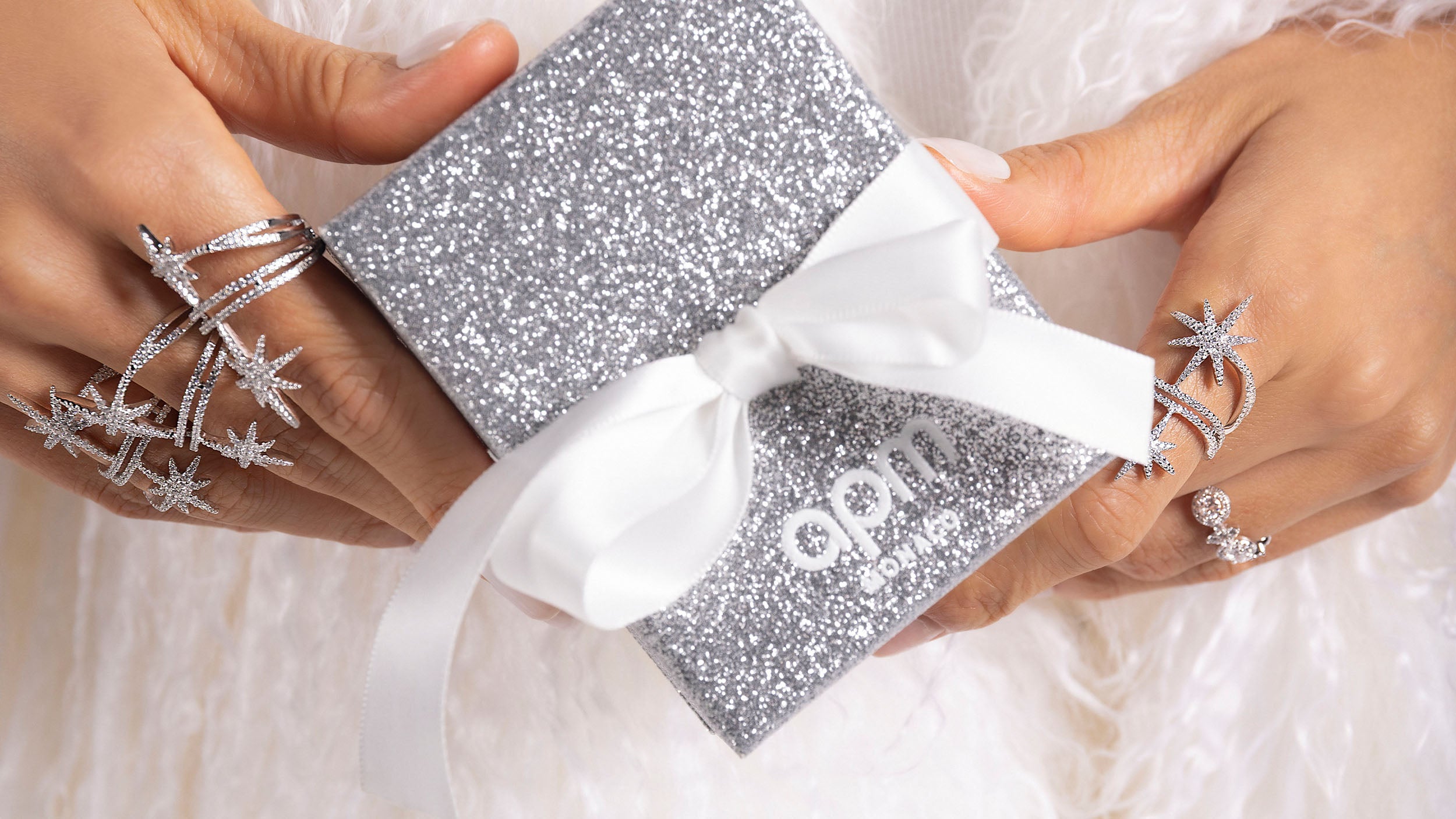 APM MONACO silver glittery box with a white bow held by hands wearing star-shaped silver rings on a light background